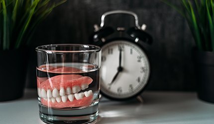 Dentures in a glass of clear fluid on a table next to an alarm clock