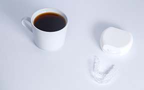 Clear aligners and their white case next to a cup of coffee on a white surface