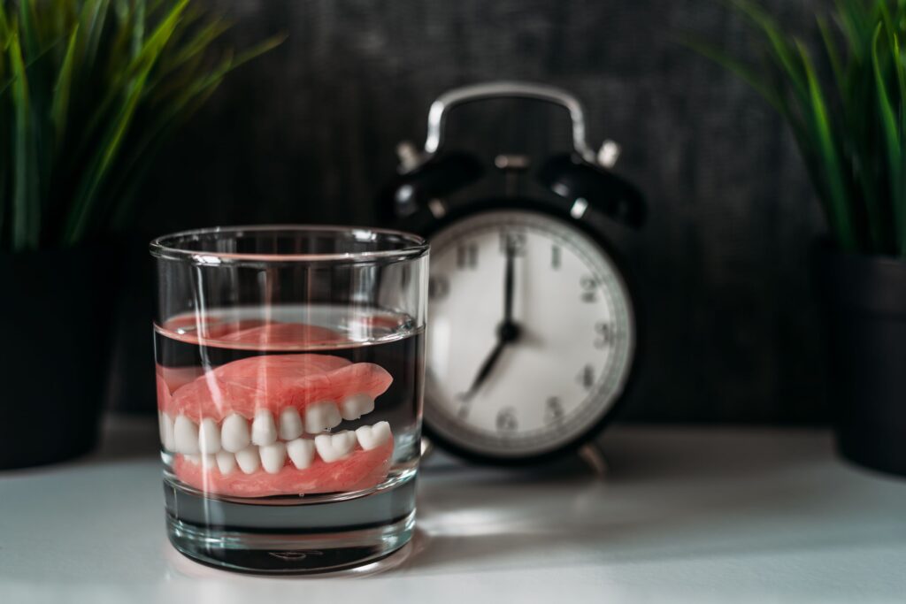 Dentures in a glass of clear fluid next to traditional alarm clock